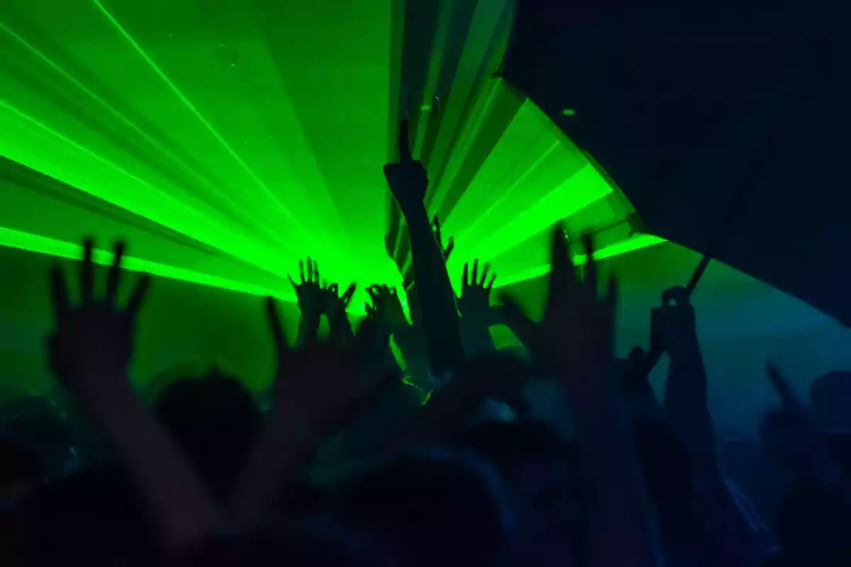 Personas bailando con los brazos al are en el interior de una discoteca con luces verdes de neón